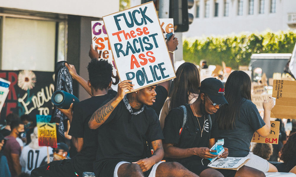un gruppo di ragazzi manifesta in America. In primo piano un ragazzo di colore alza il cartello fuck these racist ass police