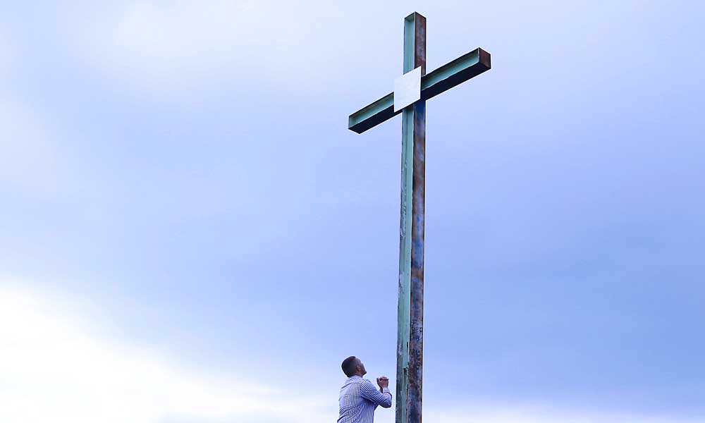 un uomo di colore prega davanti a una croce altissima, dietro un cielo pplumbeo
